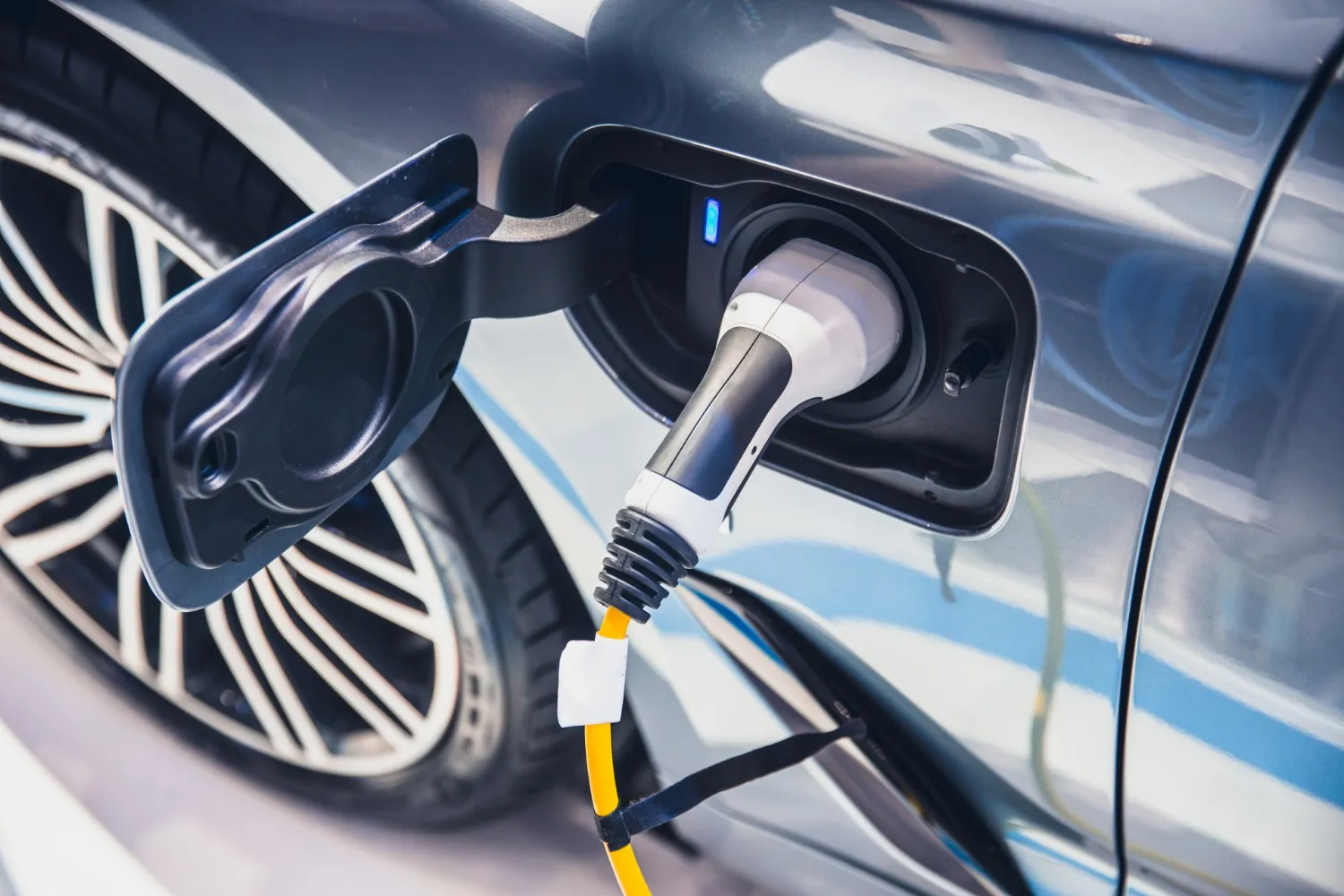 Close-up of a white and black EV charging cable plugged into a gray electric car's charging port with a yellow power cord.