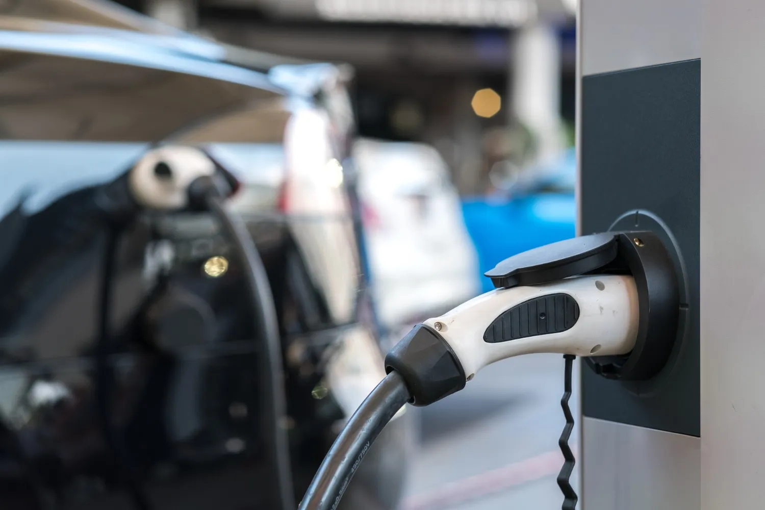 A close-up of a white electric vehicle charging nozzle plugged into a charging station with cars blurred in the background.