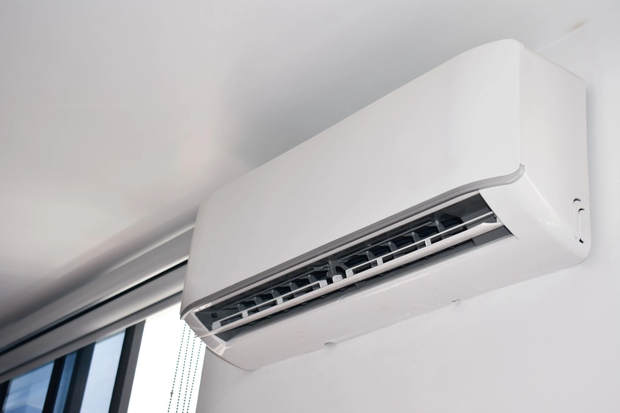 Wall-mounted white split AC unit above window with vertical blinds; vents visible for air distribution.