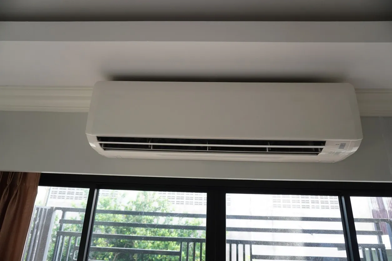 Wall-mounted split air conditioner above large black-framed windows with outdoor view of greenery and building.