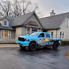 A blue pickup truck with business branding is parked on a driveway in front of a house.