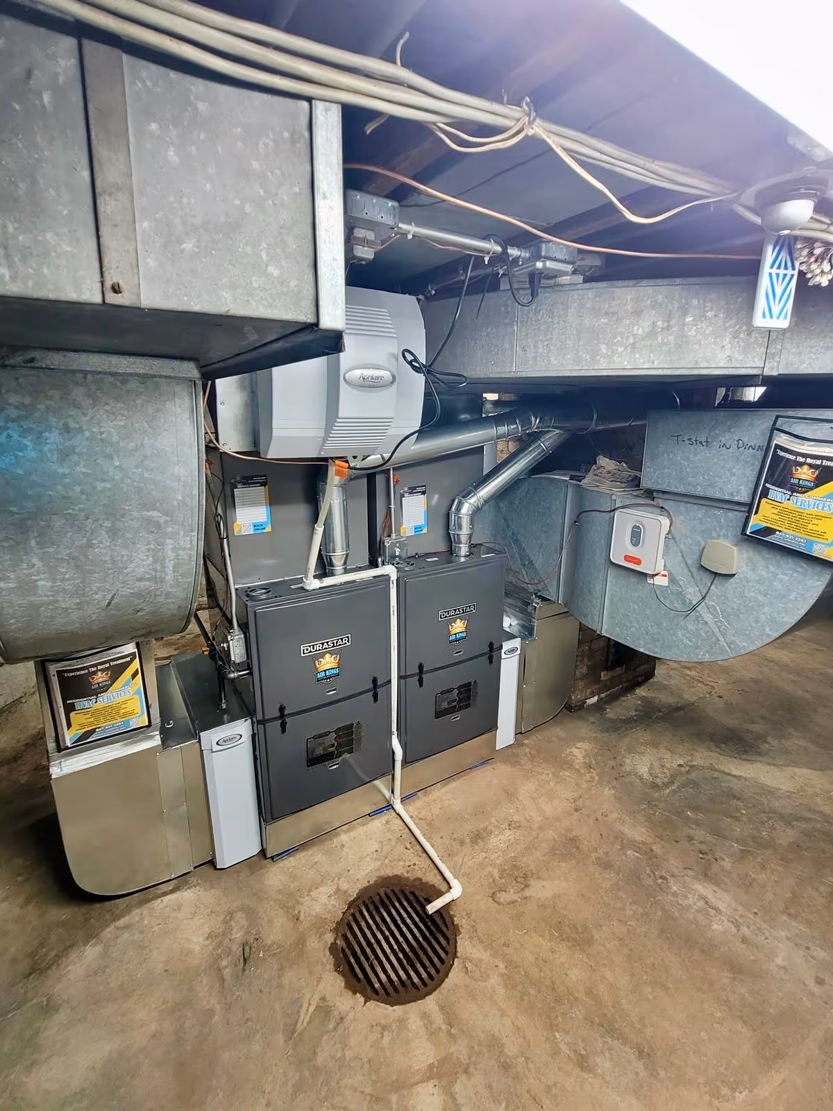 Two large HVAC units in a basement with metal ducts and a floor drain nearby.