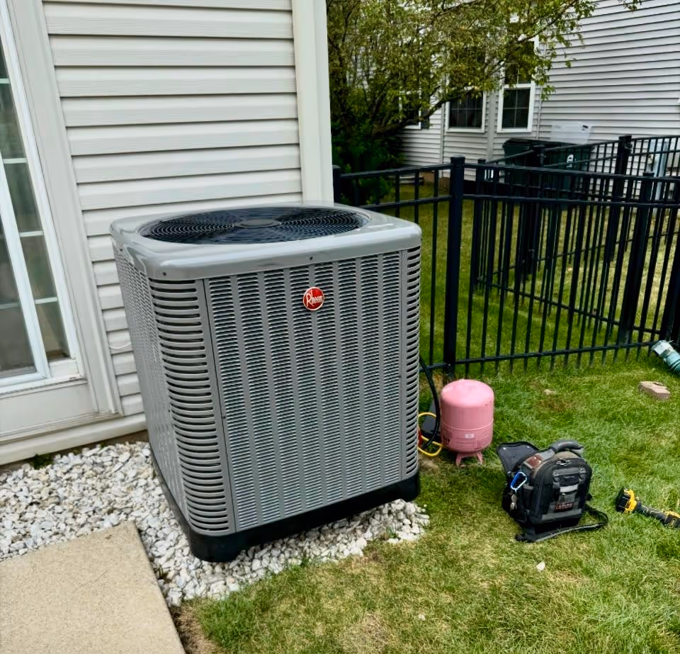 A large air conditioning unit sits outside a house near a black fence on a grassy area.