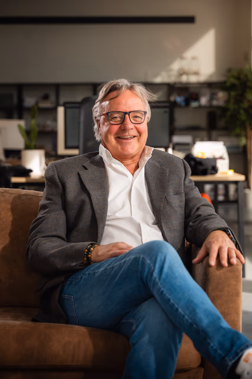 Teamlid van ’s Graevenhuys zit op een bruine zetel in een modern kantoor, glimlacht en kijkt recht in de camera, draagt witte blouse en grijze kostuumvest.