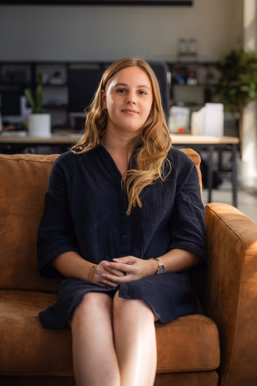 Teamlid van ’s Graevenhuys zit op een bruine zetel in een modern kantoor, glimlacht en kijkt recht in de camera, draagt een zwarte jurk.