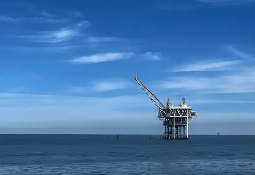 Offshore oil rig standing in calm ocean waters under a blue sky with scattered clouds.