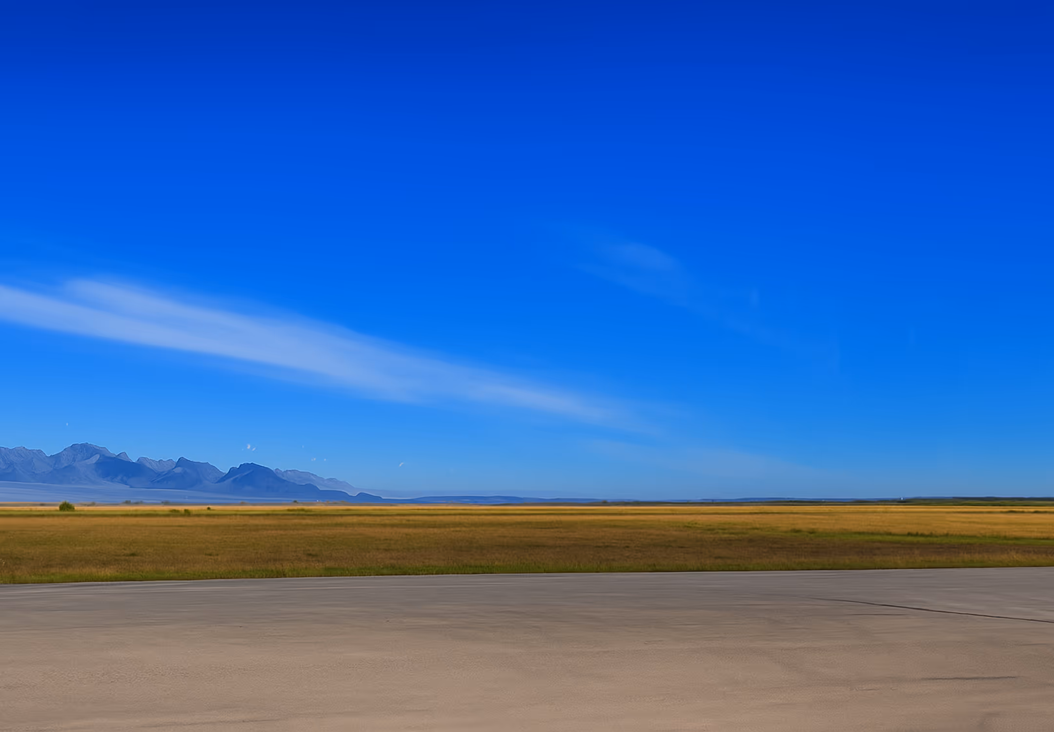 Clear blue sky over an expansive grassy field with distant mountains and a concrete foreground.