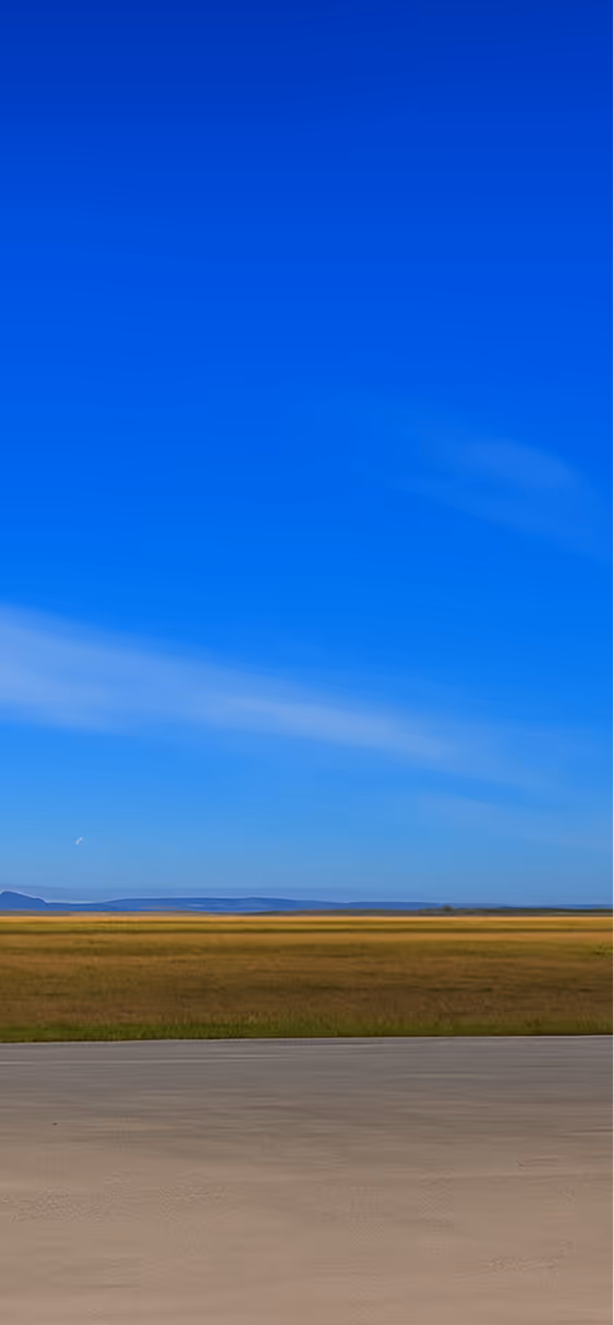 Clear blue sky over a wide open field with distant hills on the horizon and a concrete or paved surface in the foreground.