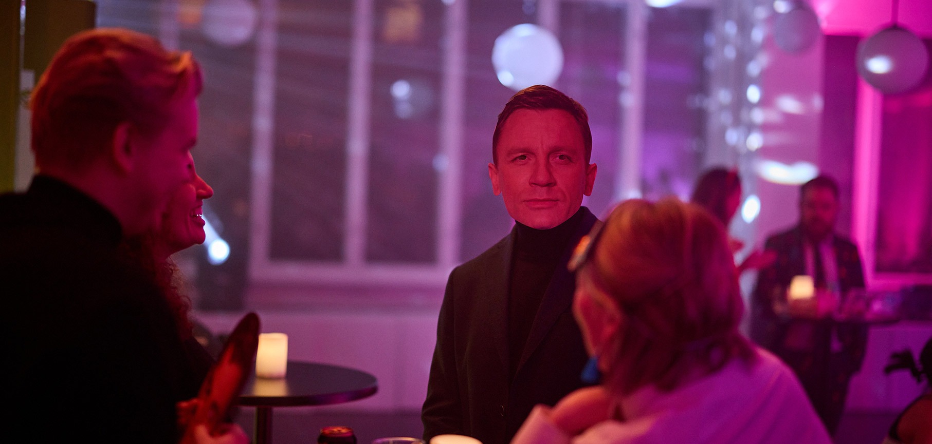 Man in black turtleneck coat looking forward at a dimly lit party with purple and pink lights, surrounded by other people socializing.