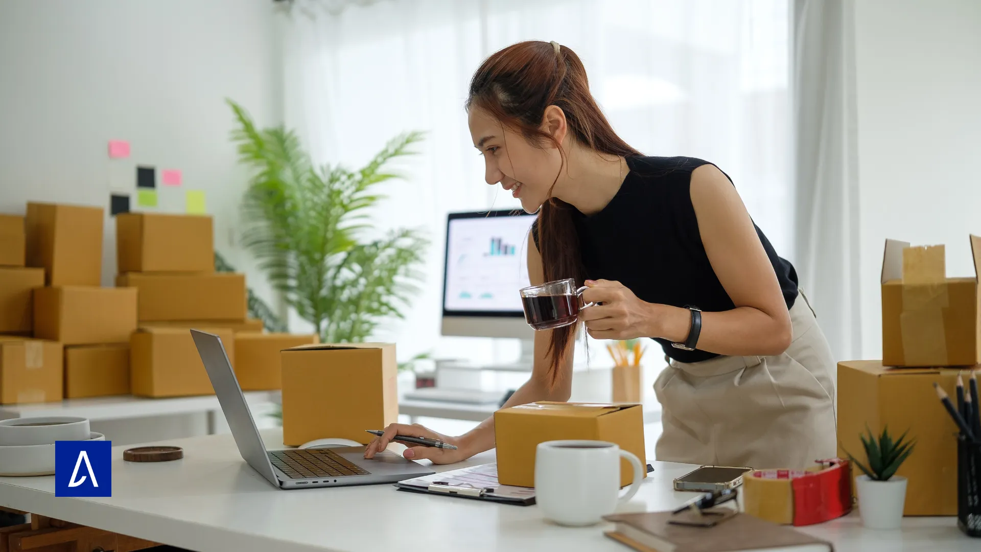 Jeune femme entrepreneure tenant une tasse de café tout en travaillant sur son ordinateur portable entourée de cartons de colis.