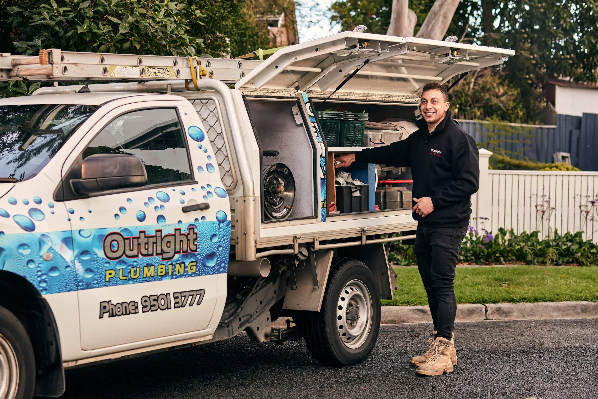 Plumber standing next to a white truck with Outright Plumbing branding, accessing tools in the truck's open side compartment.