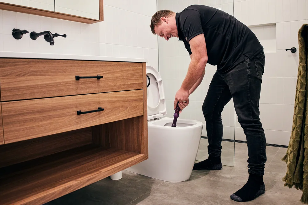Person cleaning toilet with plunger in modern bathroom with wooden vanity