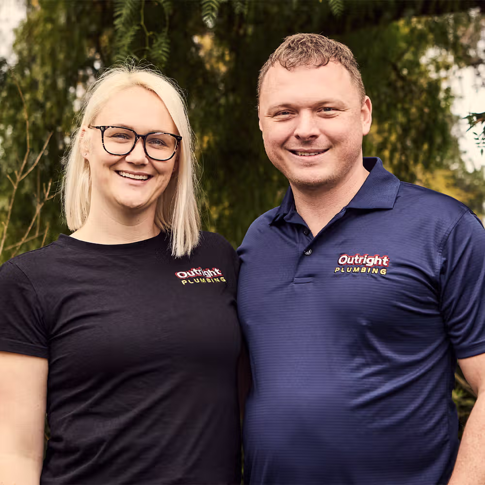 Two Outright Plumbing employees smiling in front of green foliage