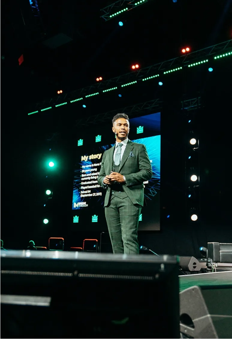Man in a green suit speaking on stage with a presentation screen behind him showing a slide titled 'My story'.