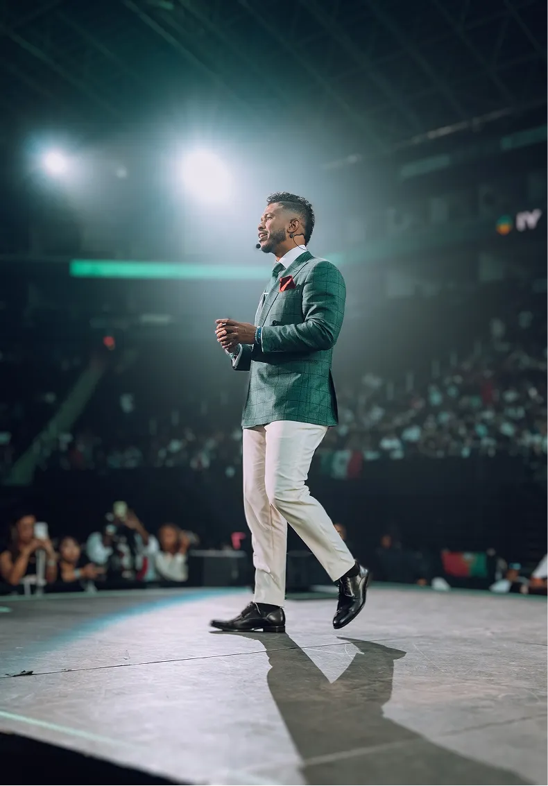 Man wearing a green blazer and white pants speaking on stage with a microphone headset in front of an audience.
