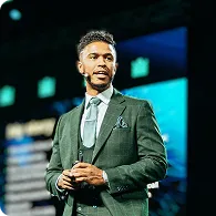 Man in a green suit and tie speaking on stage with a microphone headset.