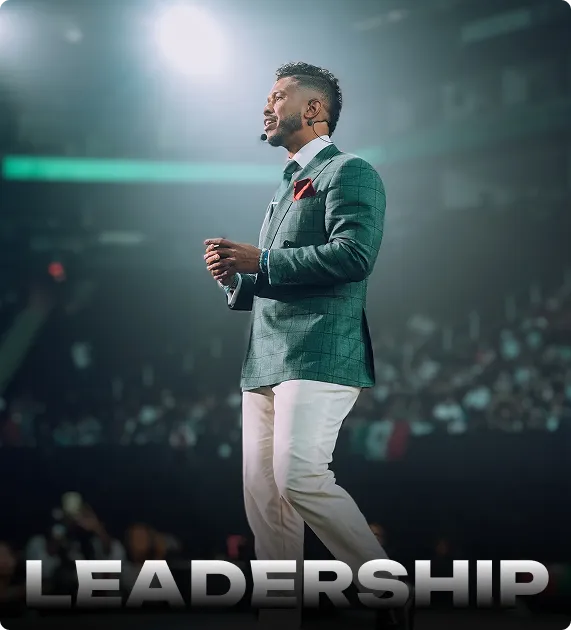Man in a green checked blazer and white pants speaking on stage with a microphone headset, with the word 'LEADERSHIP' at the bottom.