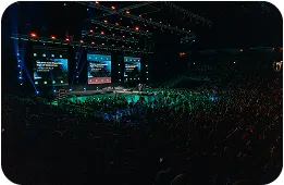 Large crowd watching a live concert or event on a stage with illuminated screens and overhead lights in a dark indoor venue.