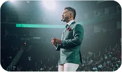 Man in a green blazer and white pants speaking on stage in front of a large audience.