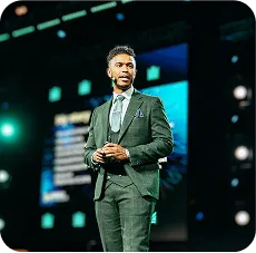 Man in a dark suit and tie speaking on stage with a large screen in the background.