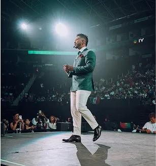 Man in a gray blazer and white pants speaking on stage in front of a large audience in an indoor arena.