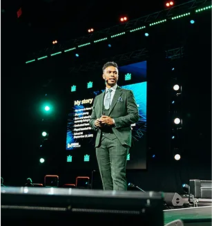 Man in a gray suit speaking on stage with a presentation screen behind him.