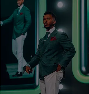 Man in a dark green suit jacket and white pants standing on stage with a microphone headset and red pocket square.