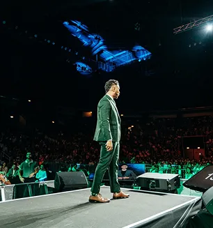 Man in green suit speaking on stage in front of a large audience with blue light installation above.