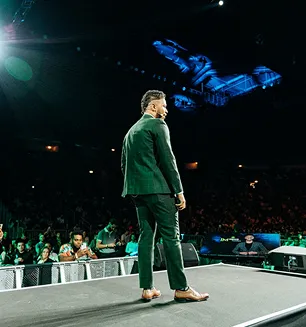 Man in green suit standing on stage facing a large audience with a blue digital graphic projected behind him.