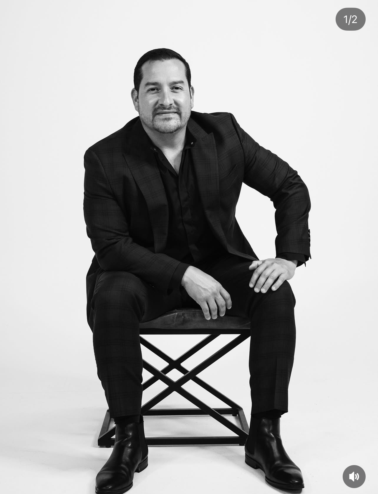 Man in a dark suit sitting on a chair against a plain white background, looking at the camera.
