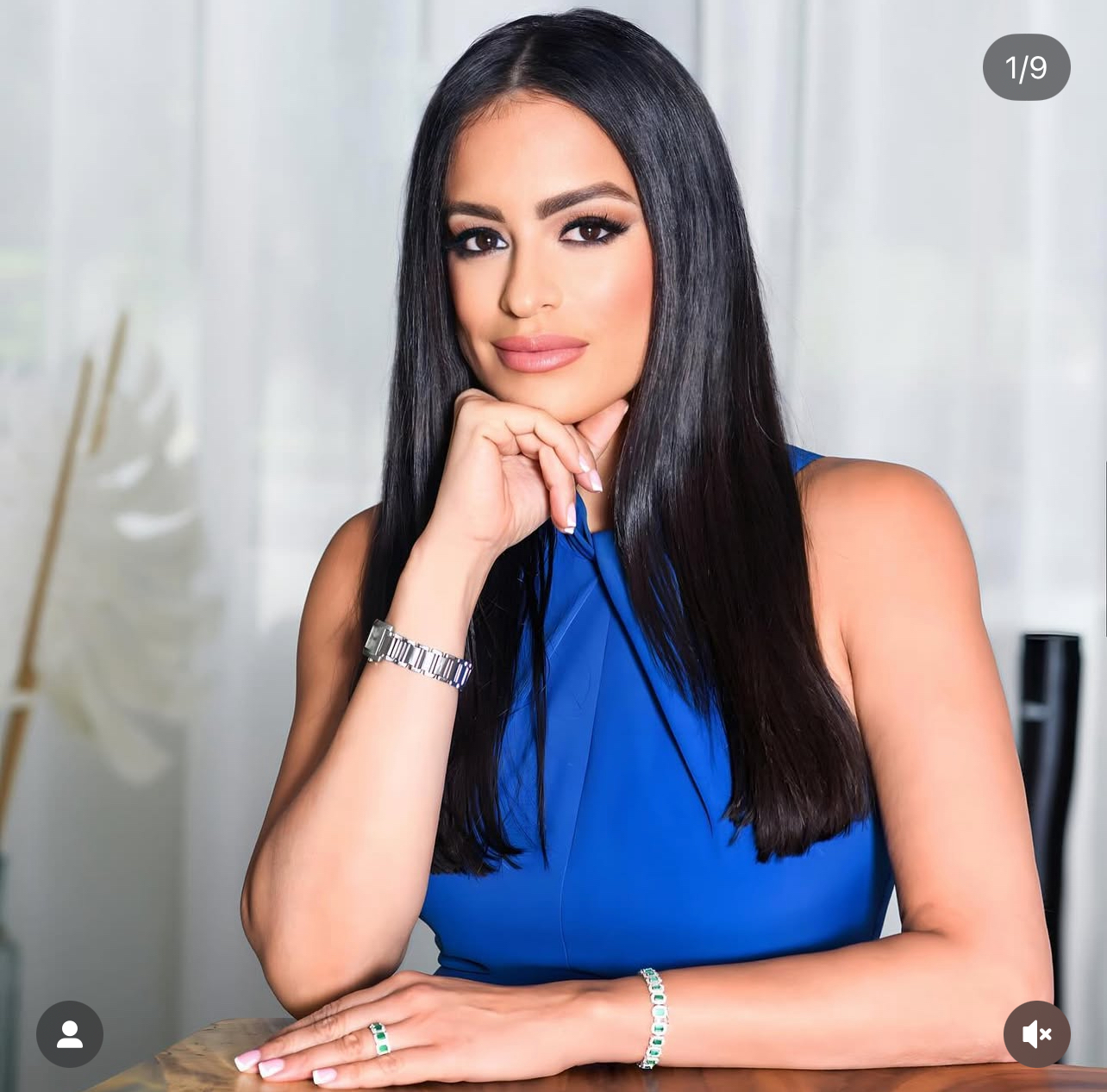 Woman with long black hair wearing a blue sleeveless top, resting her chin on her hand at a table.