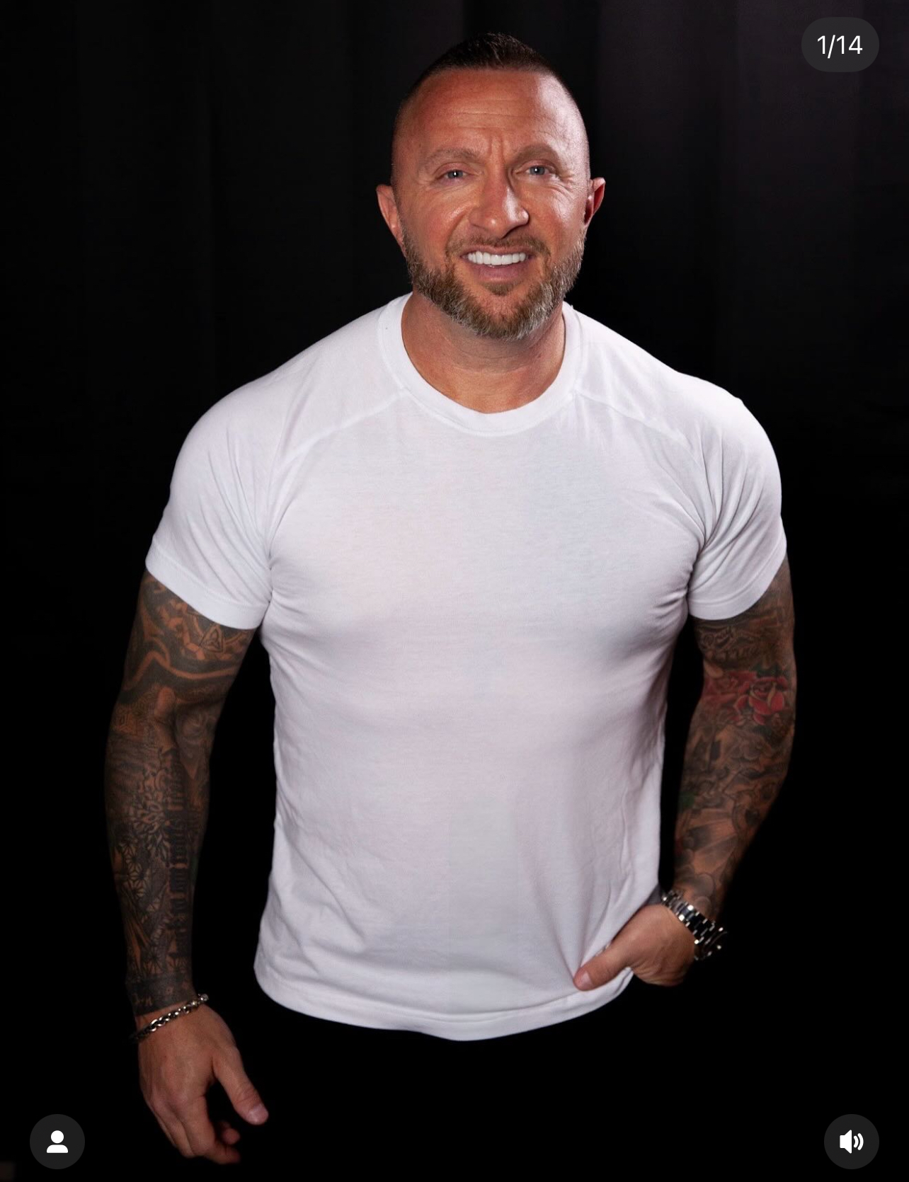 Smiling man with short hair, a beard, and full sleeve tattoos wearing a fitted white t-shirt against a black background.