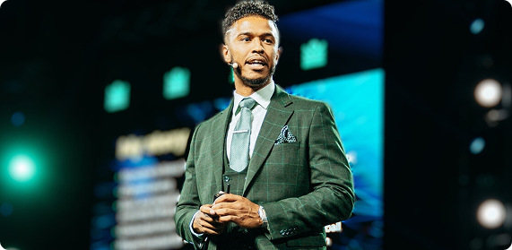Man in a green plaid suit speaking on stage with a headset microphone.