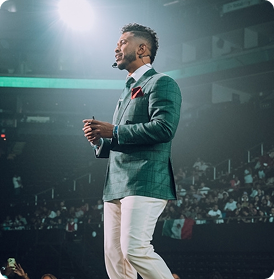 Man in a checkered blazer and white pants speaking on stage with a headset microphone in front of a large audience.