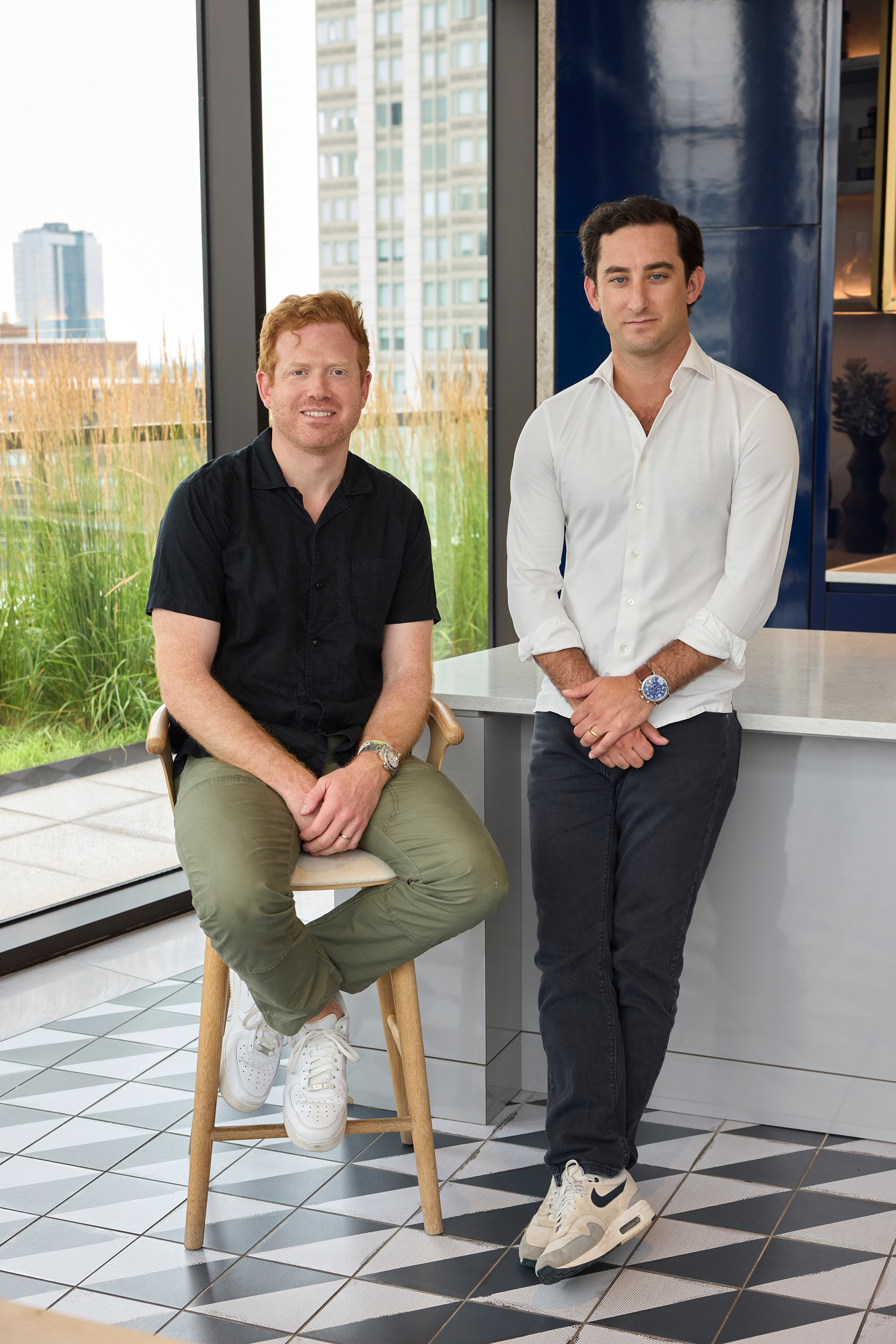Tom and Brian from The Story Shack - Brian sitting on a wooden stool and Tom standing beside a countertop, with large windows and tall grasses visible in the background
