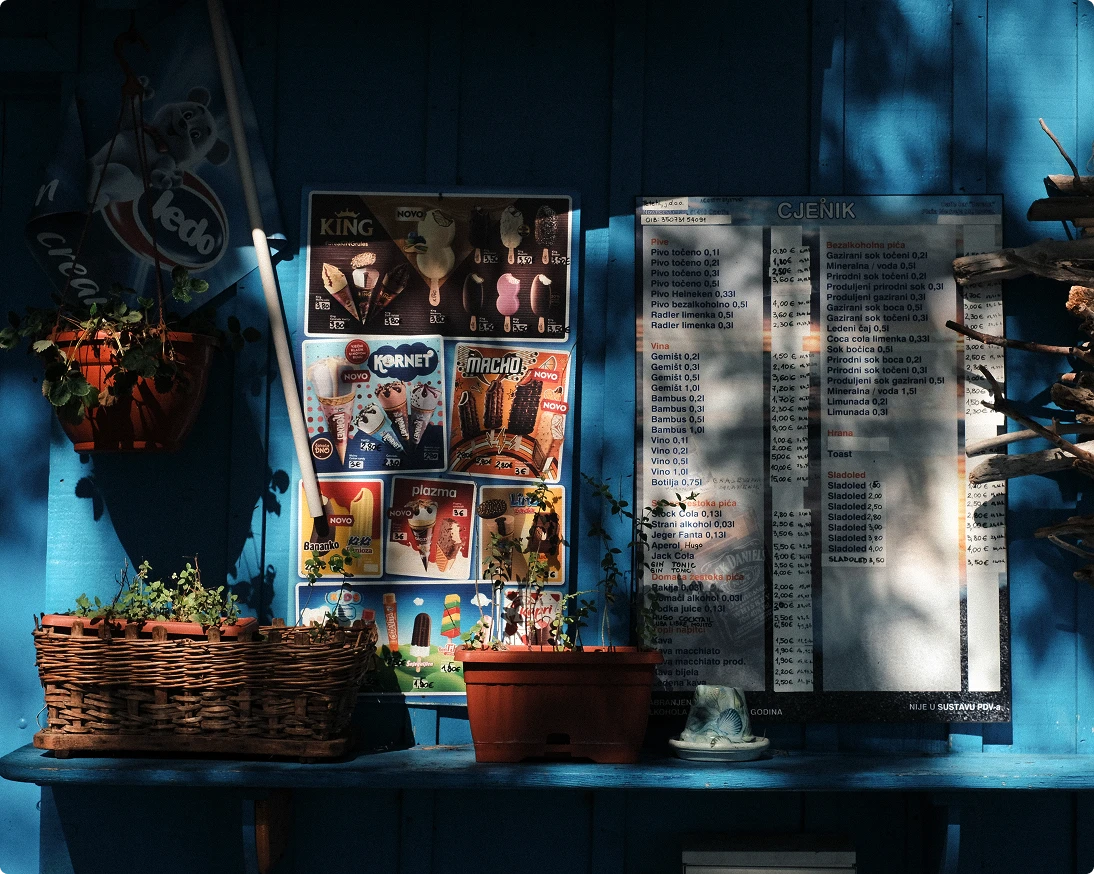 Sunlit blue wall displaying ice cream and beverage menu boards with potted plants and wicker basket on a wooden shelf.