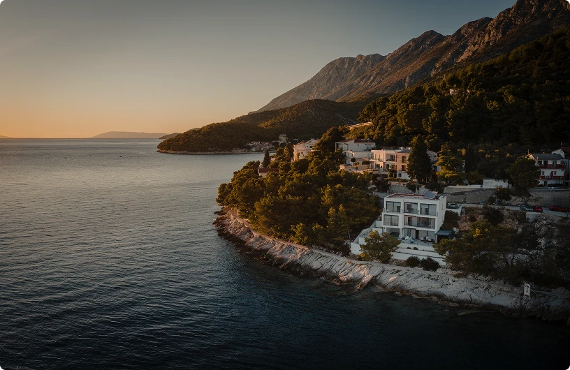 Coastal village with white houses nestled among pine trees on rocky shoreline at sunset, with mountains in the background.