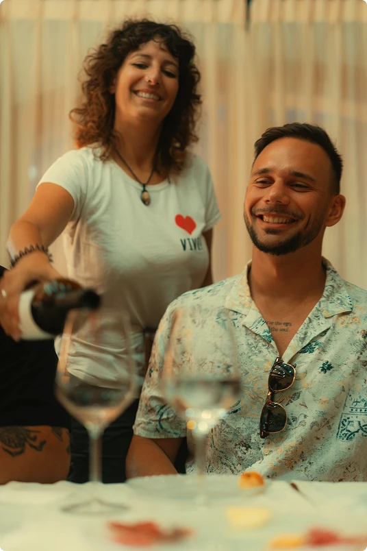 Smiling woman pouring wine into a glass for a smiling man seated at a table.