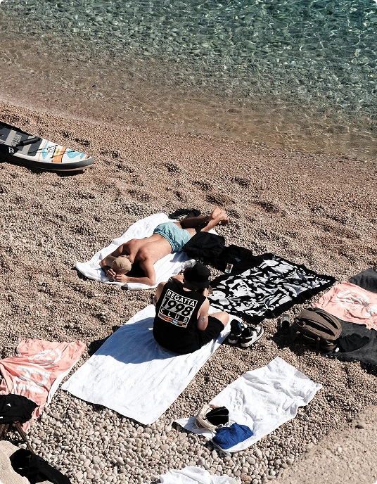 Two people relaxing on towels on a pebble beach next to clear turquoise water, with one person lying down and the other sitting.