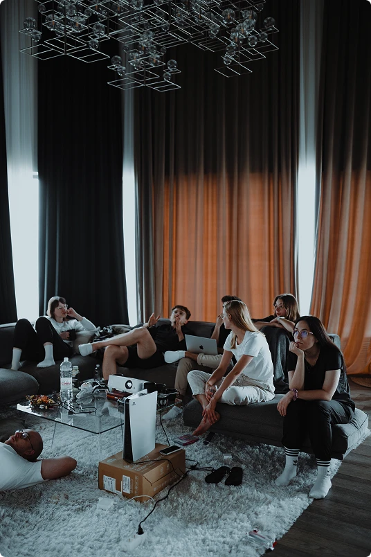 Group of six young adults sitting and lounging on sofas in a cozy living room with dark curtains, a projector, and a laptop.