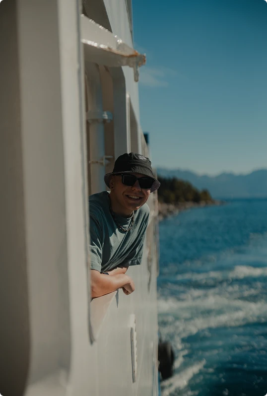Young person wearing a bucket hat and sunglasses leaning out of a boat window smiling with water and distant mountains in the background.