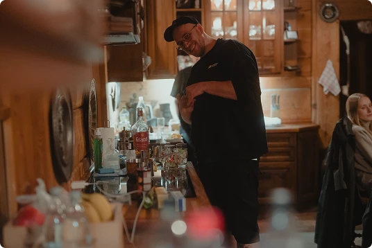Man wearing a black cap and glasses smiling behind a kitchen counter with various bottles and glasses.