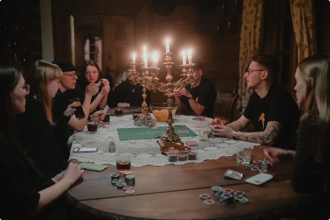 Group of people sitting around a table playing cards, illuminated by ornate candelabras with lit candles.