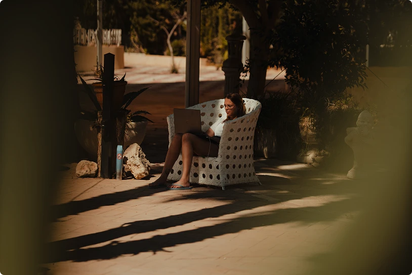 Woman sitting in a woven chair outdoors, working on a laptop in dappled sunlight.