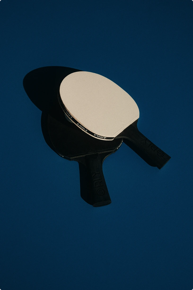 Two table tennis paddles, one with a white rubber side facing up, lying on a dark blue surface casting shadows.