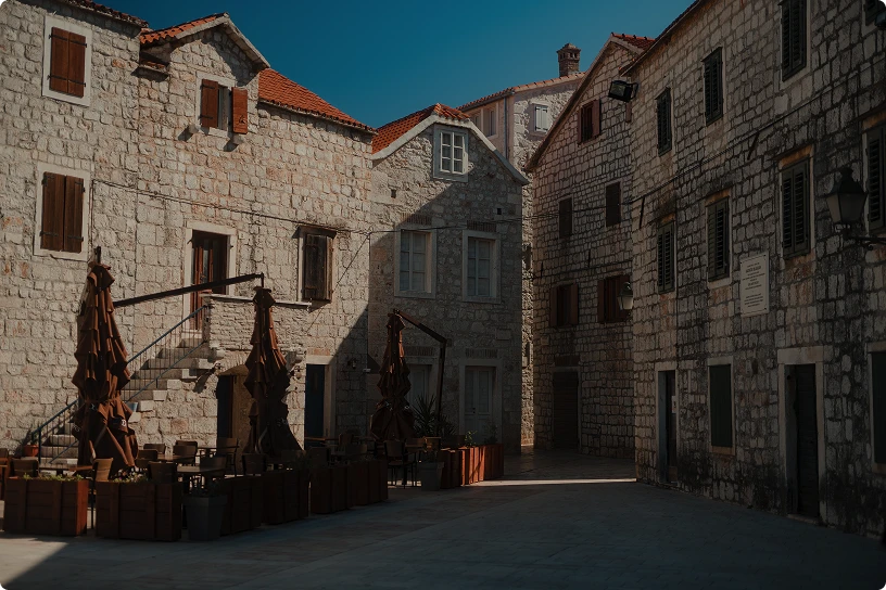 Sunlit narrow street with stone buildings and closed umbrellas shading outdoor café seating.