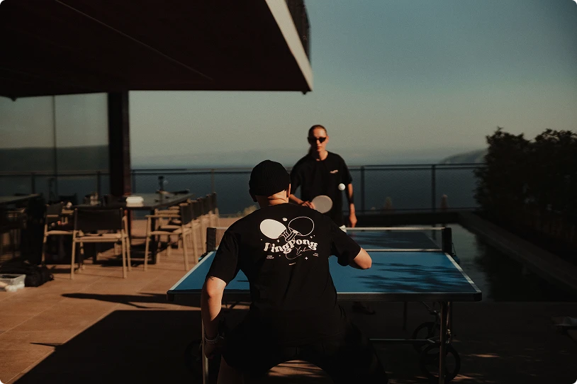 Two men playing table tennis outdoors on a terrace with ocean view during sunset.