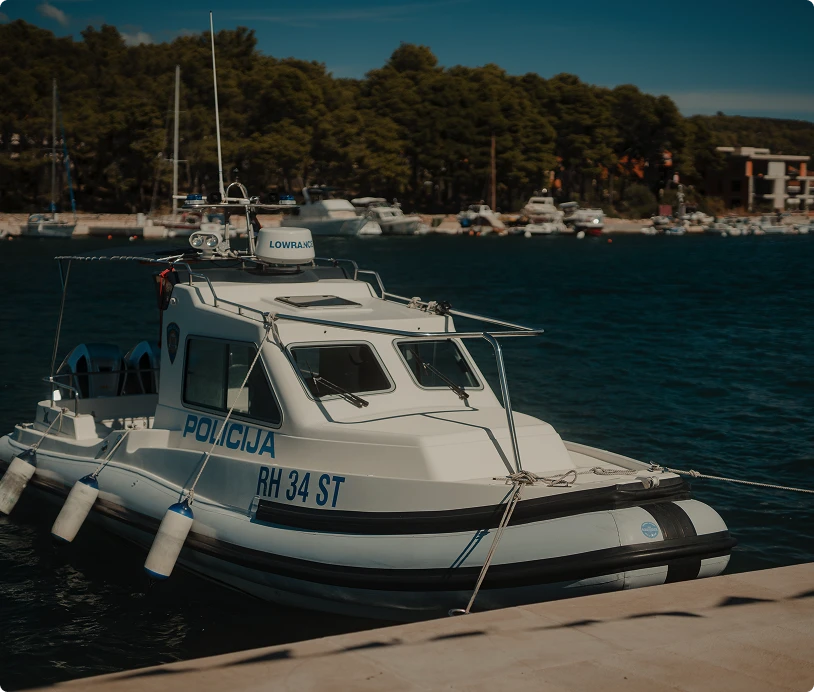 White police boat docked at a pier with 'POLICIJA RH 34 ST' written on its side and a forested shoreline in the background.