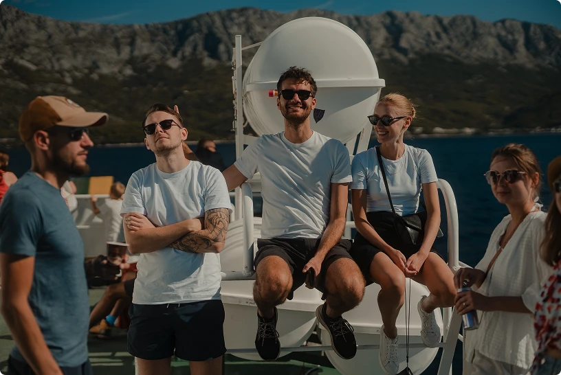 Group of six young adults wearing sunglasses relaxing on a boat with mountainous coastline and blue water in the background.
