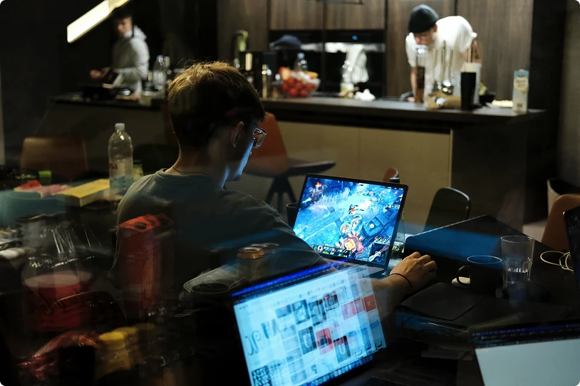 Person wearing glasses playing a video game on a laptop in a dimly lit room with other laptops and kitchen area in the background.